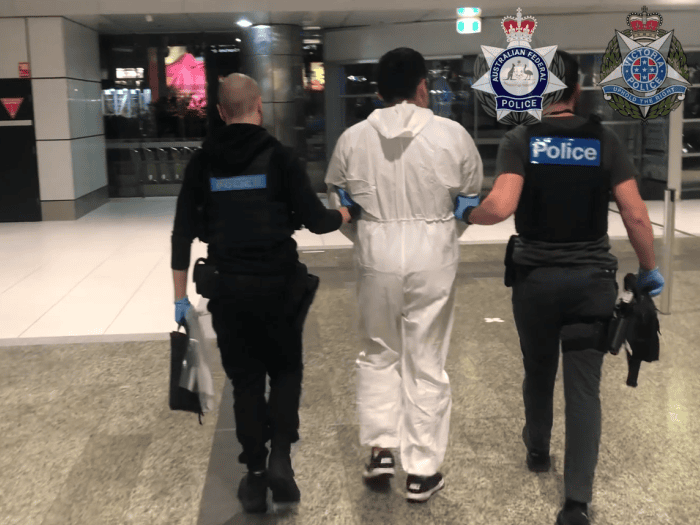 A man in a white disposable medical scrubs is led away from the camera by two police officers