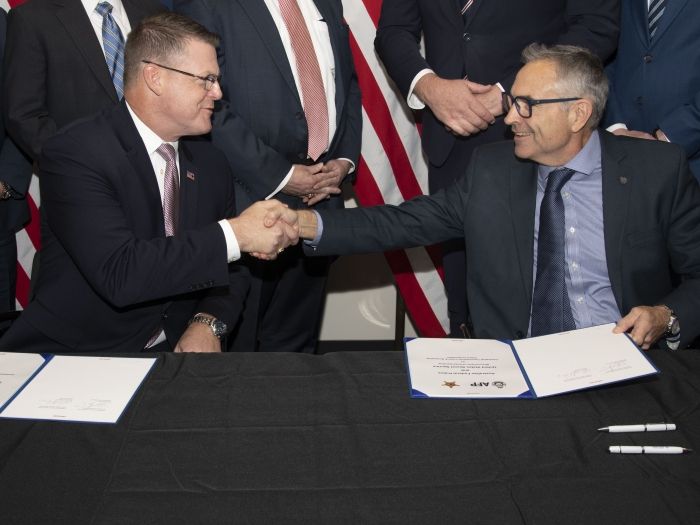 Two men seated at a black table shake hands.