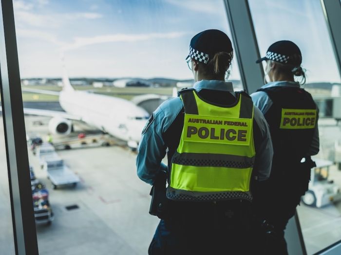 Two Australian Federal Police officers wearing checkered caps and high-visibility vests stand at an airport terminal window, overlooking an aircraft on the tarmac. Ground service vehicles can be seen near the plane.