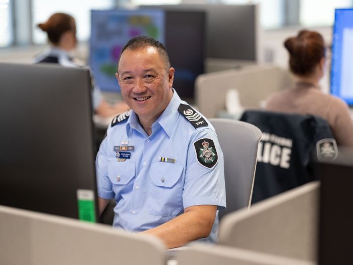A smiling man in an AFP police officer uniform sits at a desk with a large monitor and 2 other staff sit behind him facing their computer monitors.