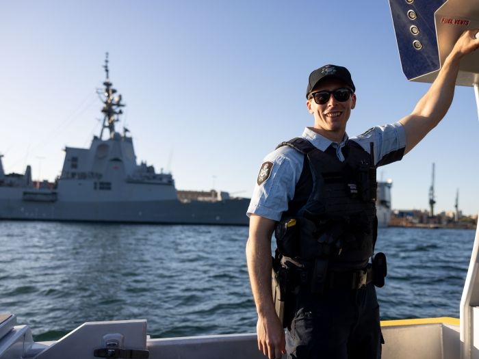 Protective Service Officer on a boat in Sydney Harbour with a large naval behind him