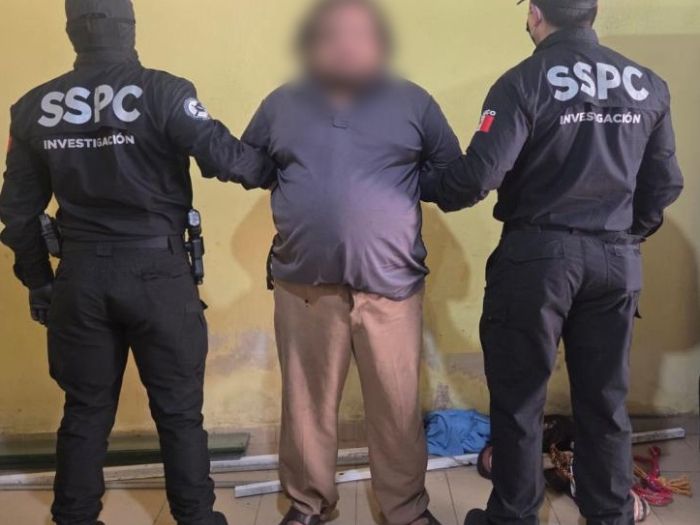 A handcuffed man facing the camera with two police officers holding him with their backs to the camera
