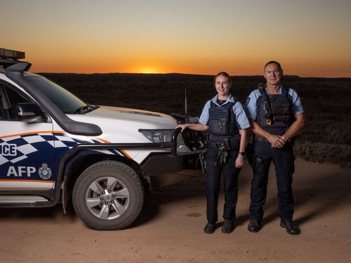 Two AFP officers standing beside a marked police four-wheel drive vehicle on a dirt road at sunset in a remote Australian landscape.