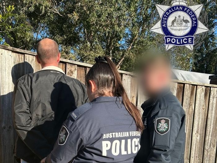 Image depicts a man in handcuff against a wooden fence with two police officers behind him
