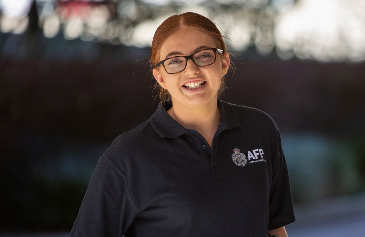 A woman with red hair pulled back, wearing glasses and an AFP (Australian Federal Police) black polo shirt, smiles at the camera. 