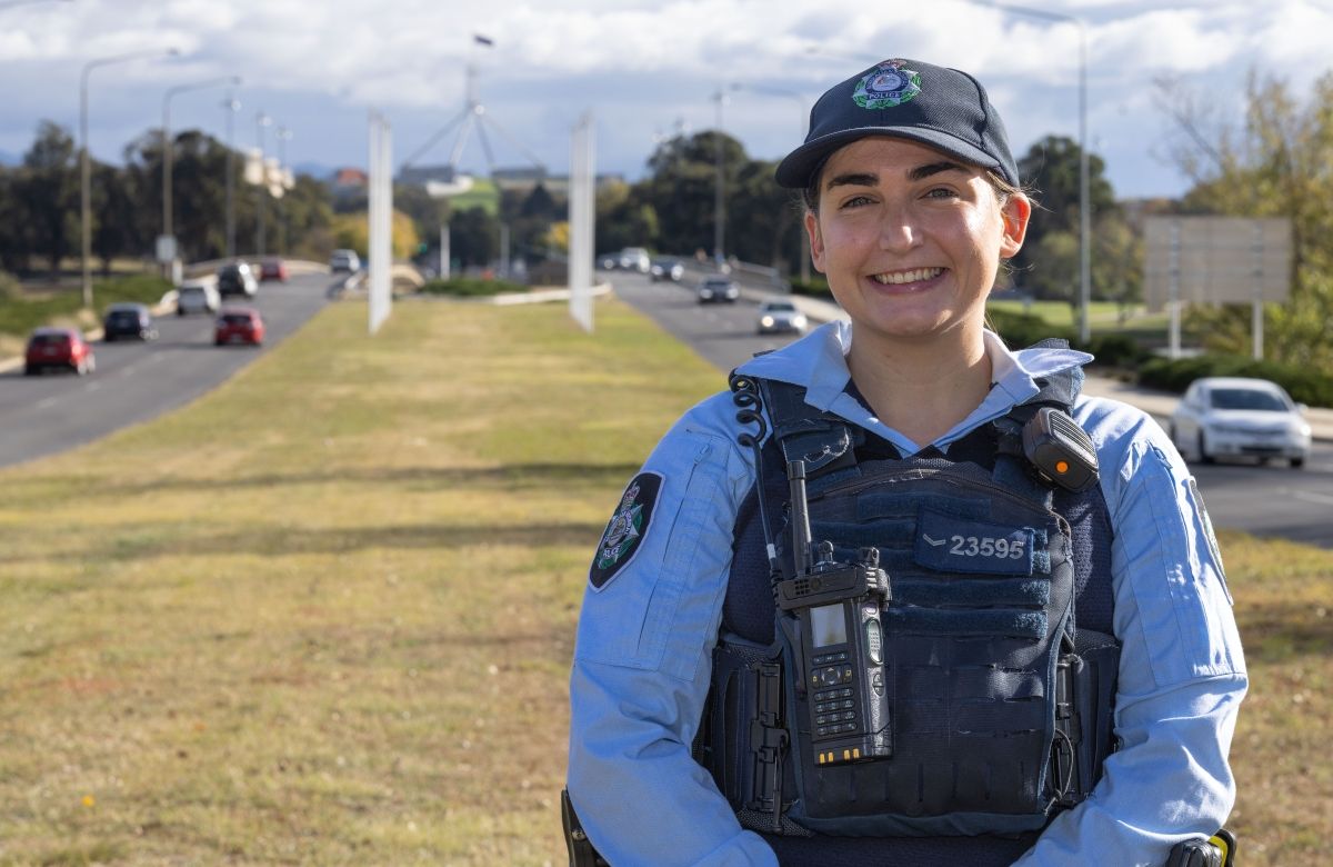 AFP Professional Service Officer standing on grass with roads either  side and Parliament House behind her.