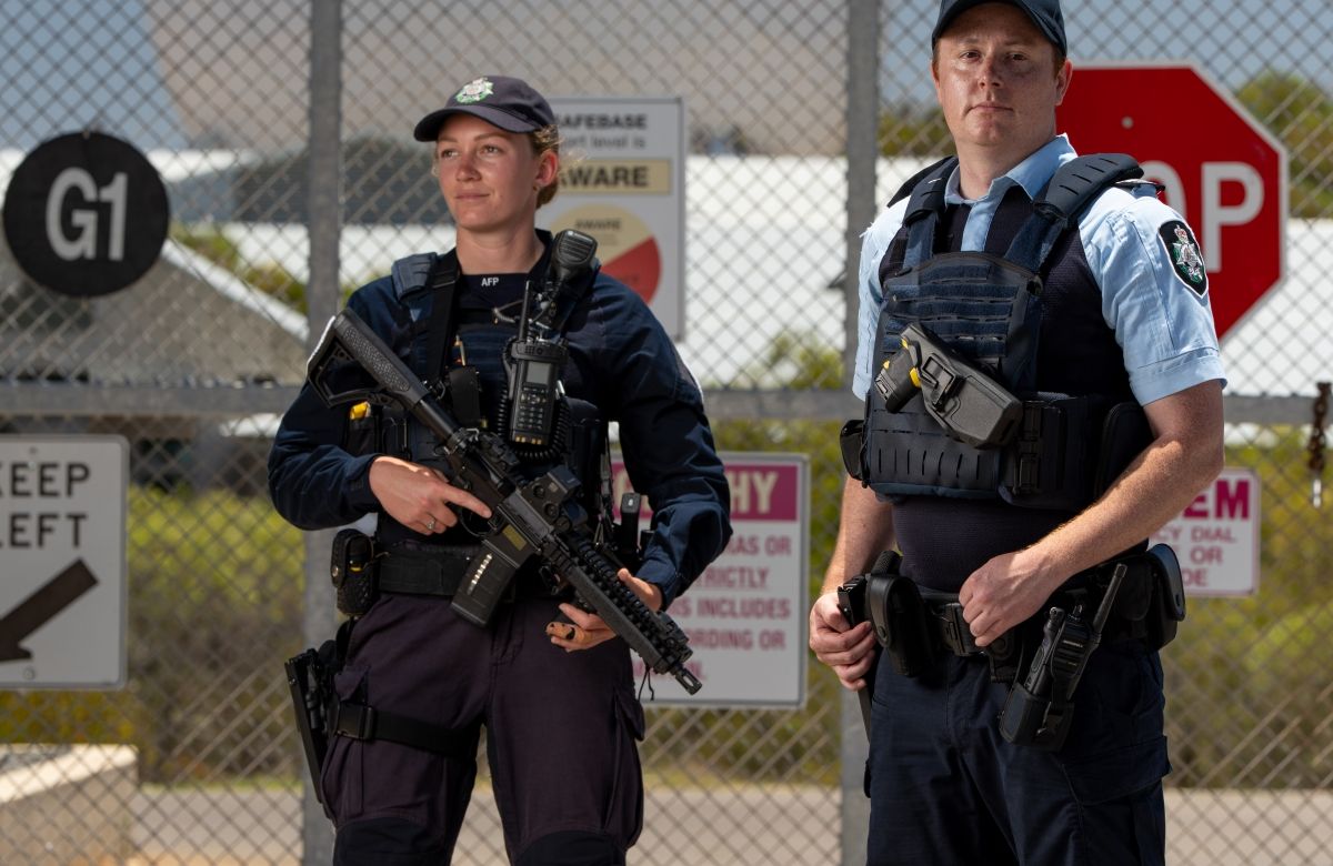 Two AFP officers standing in front of a secure facility entrance, with fencing and infrastructure visible in the background.