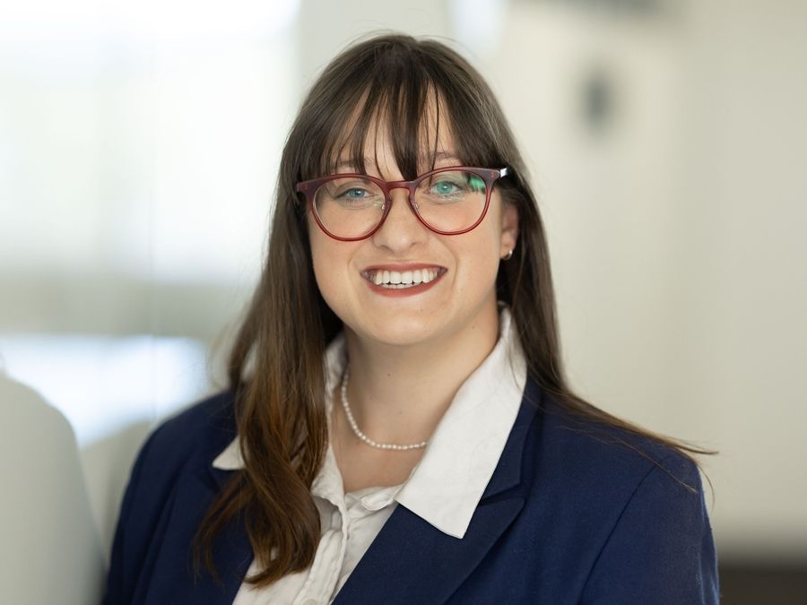 Portrait of Elizabeth. She is wearing a navy blue blazer and white shirt and has long brown hair and glasses. 
