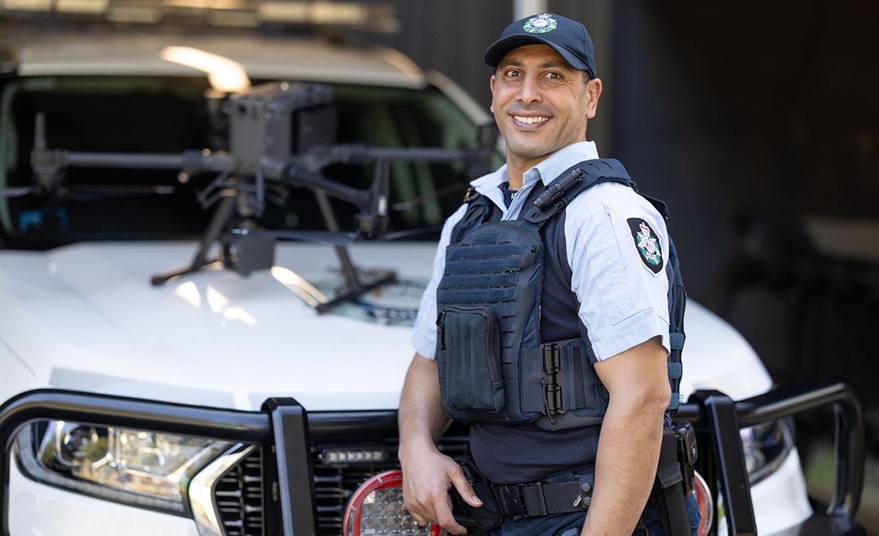 A uniformed office, with a drone on the bonnet of a white police vehicle behind him