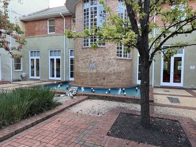 A small courtyard with a brick building, a circular tiled water feature with small fountains, paved brick paths, and a leafy tree in the foreground.