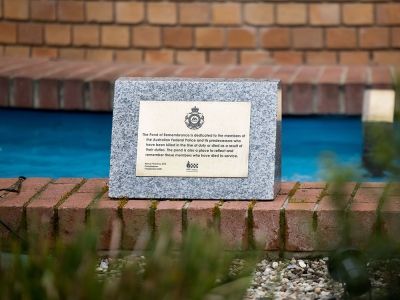 A stone memorial plaque mounted on a low brick wall beside a water feature. The plaque reads "The Pond of Remembrance is dedicated to the members of the Australian Federal Police and its predecessors who have been killed in the line of duty or died as a result of their duties. The pond is also a place to reflect and remember those members who have died in-service."