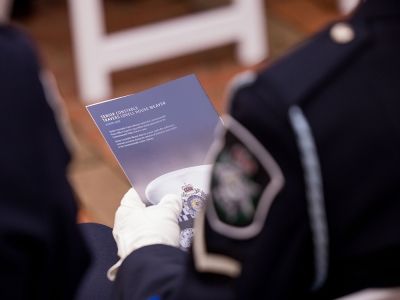 A gloved hand holds a printed memorial program featuring a police emblem, with a police uniform visible in the foreground.