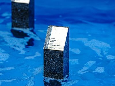 Two small granite memorial markers standing in bright blue water, with brick edging visible behind them.